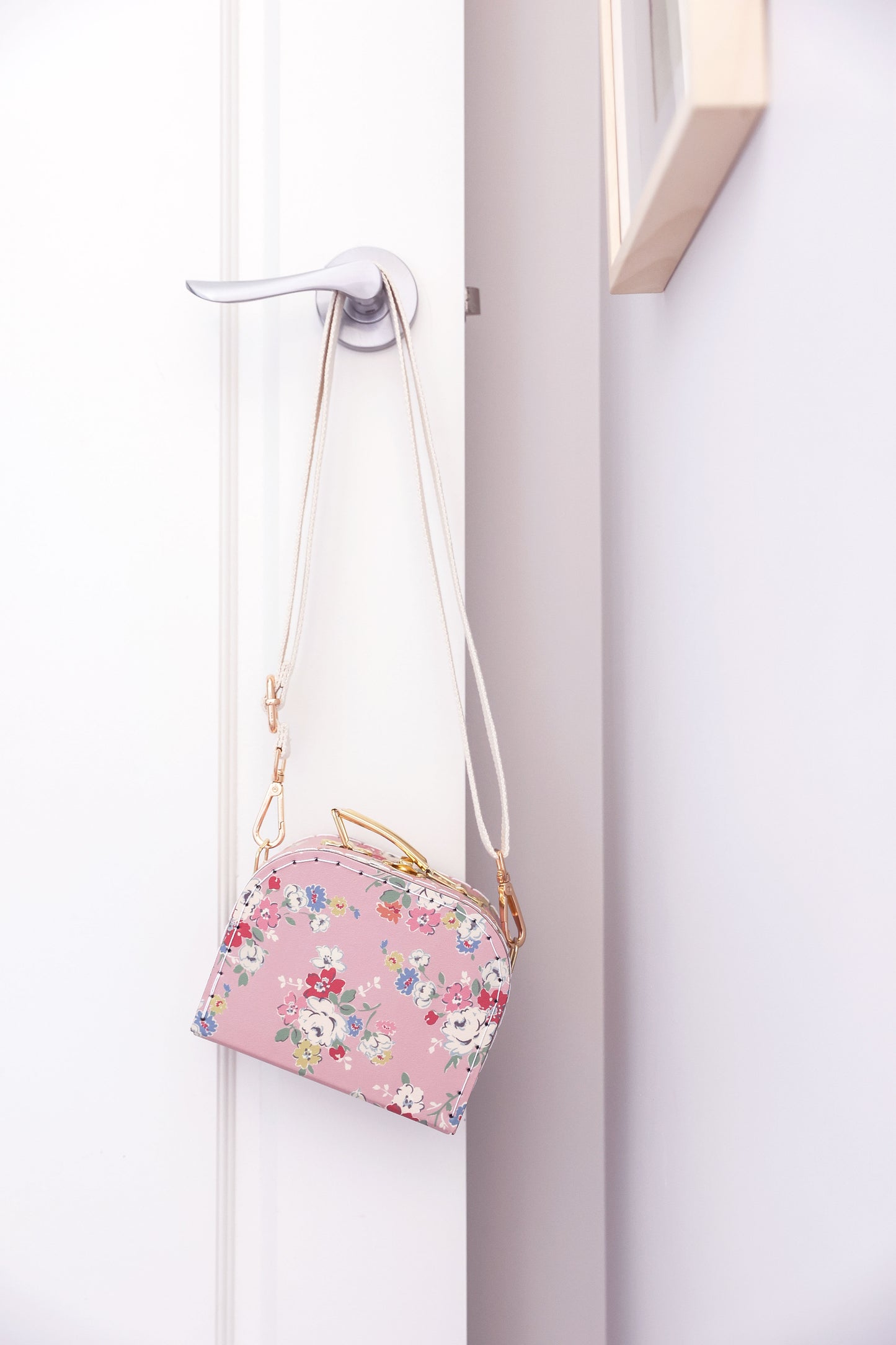Pink crossbody case with floral print and gold chain strap hanging on a door handle.
