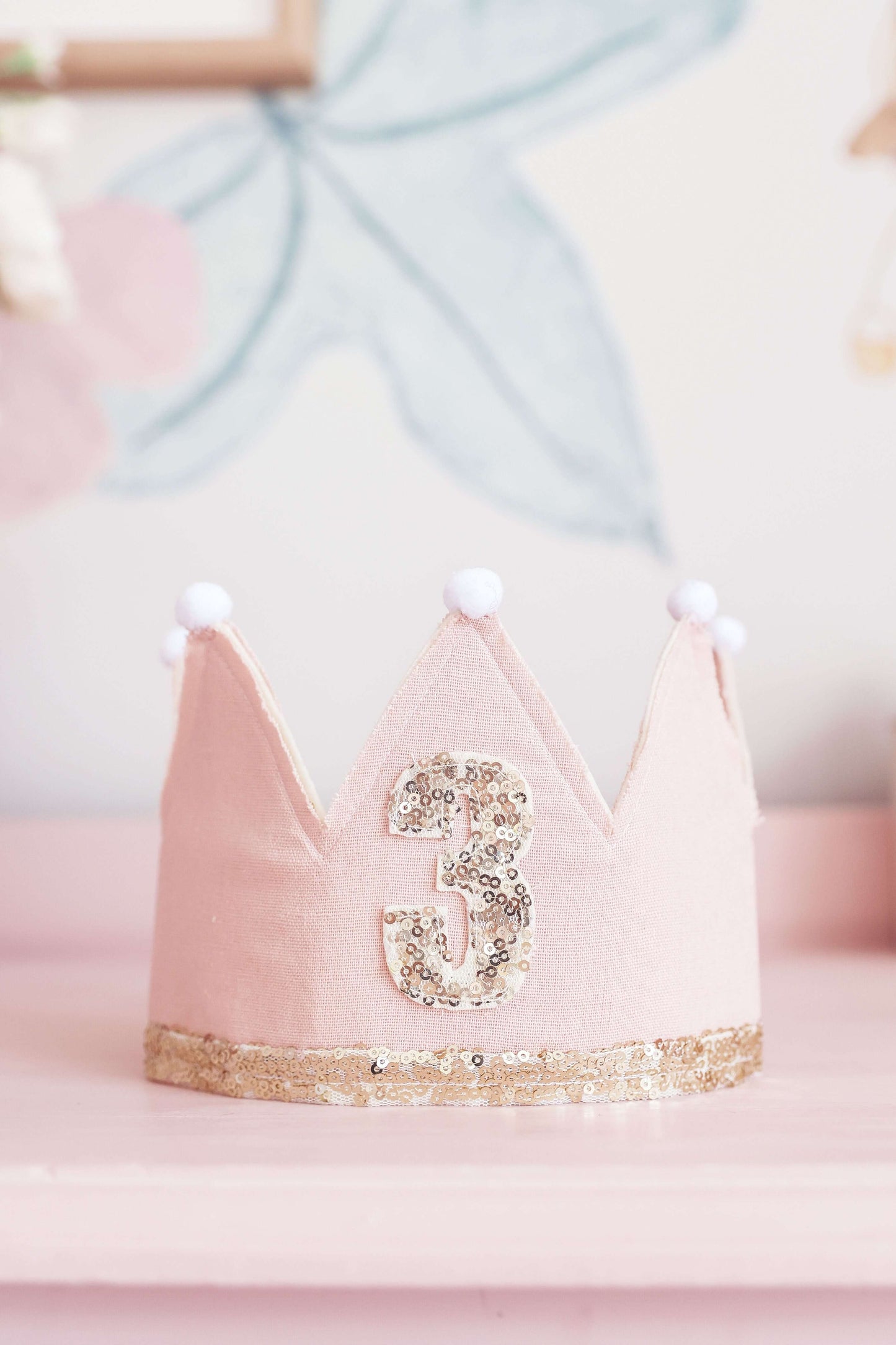 Adjustable pink crown with gold and sequin for 3rd birthday celebration, featuring pom-poms and linen backing.