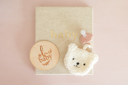 Sherpa bear pacifier comforter ivory next to baby milestone disc on soft pink background.
