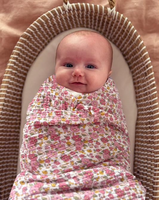 Smiling baby swaddled in pink floral muslin blanket with small rose pattern.