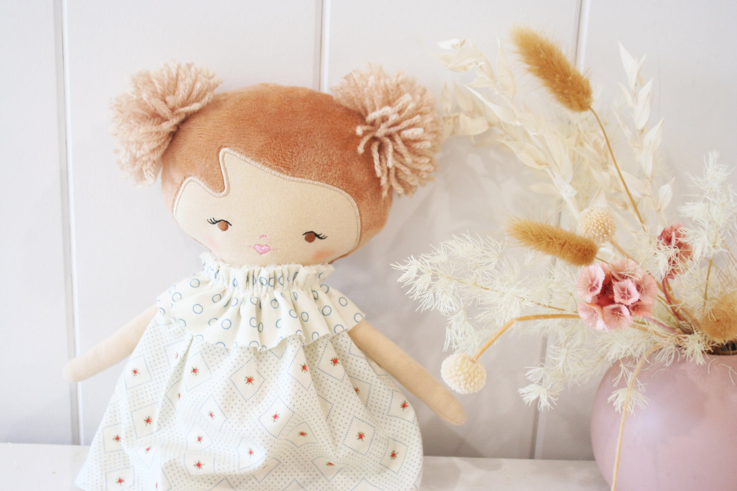 Soft fabric doll with light brown yarn hair in two buns, wearing a white dress with small orange dots and blue rose accents.