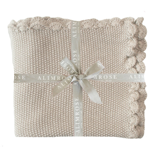Latte-colored knit baby blanket with moss stitch and scalloped edges tied with a cream ribbon.