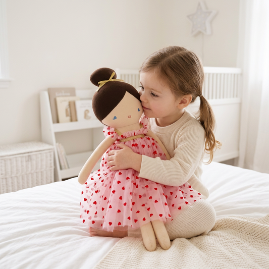 52cm soft fabric doll with brown hair bun and red heart-patterned pink dress held by toddler on bed.