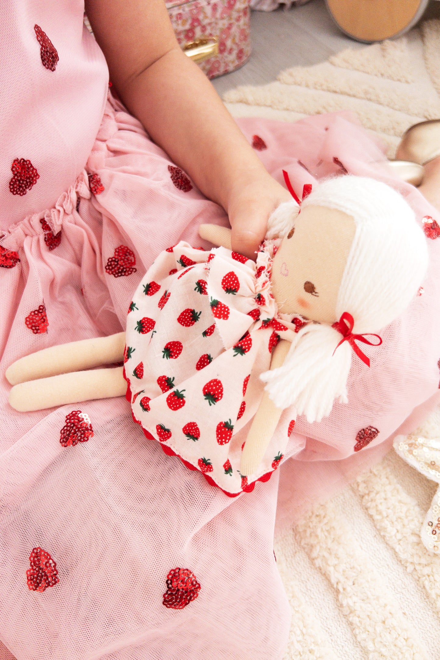Cloth doll with white braided hair in a red strawberry-print dress held by a child in a pink dress with red heart appliques.