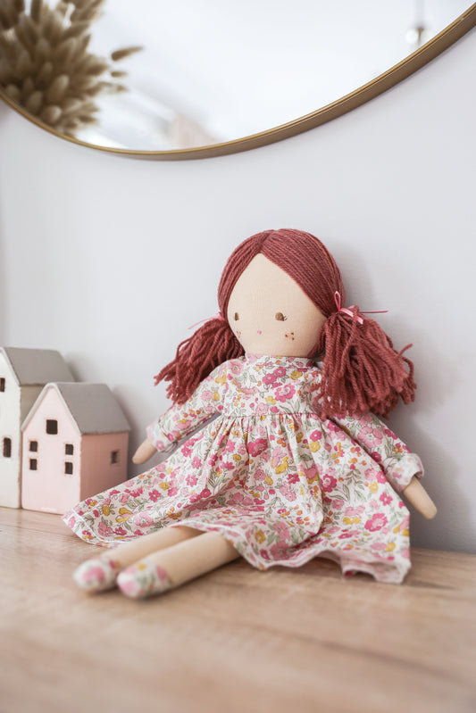 Soft fabric doll with brown yarn hair in pigtails, wearing a floral pink and white dress, seated on a wooden surface.