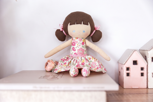 Soft cloth doll with brown yarn hair in pigtails, wearing a pink floral dress and matching shoes, sitting on a book next to.