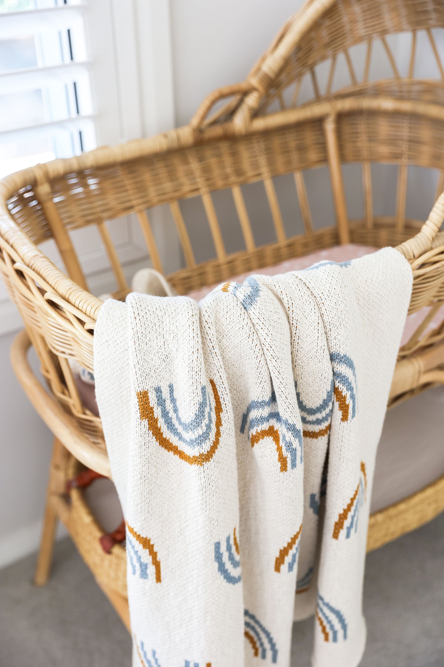 White baby blanket with soft texture and rainbow arches in muted blue, orange, and gray draped over a woven bassinet.