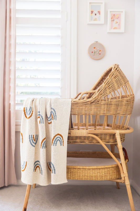 Soft cream baby blanket with pastel rainbow patterns draped over a wicker bassinet in a nursery setting.
