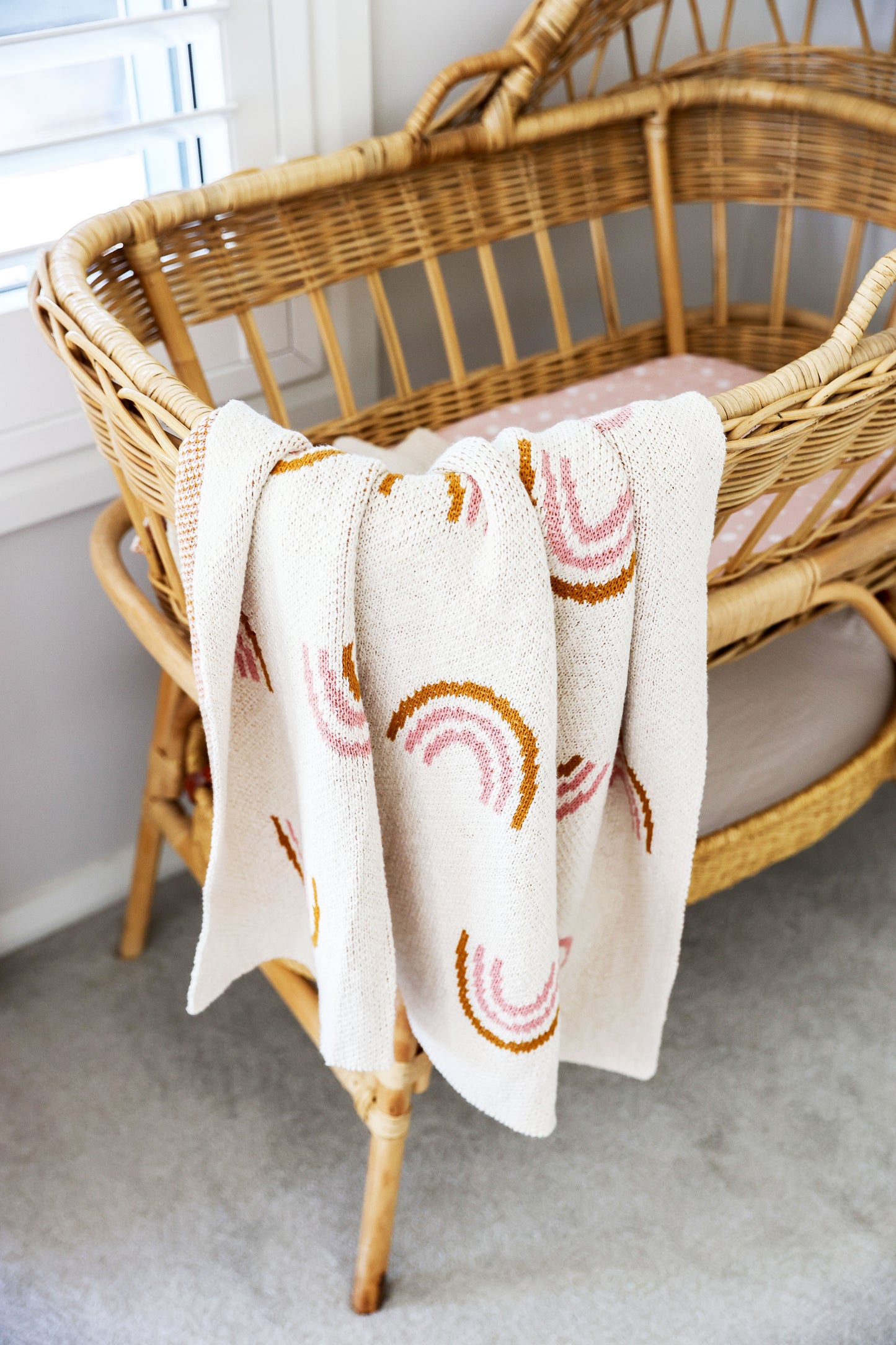 Soft white baby blanket with pink and orange rainbow patterns draped over a wicker crib.