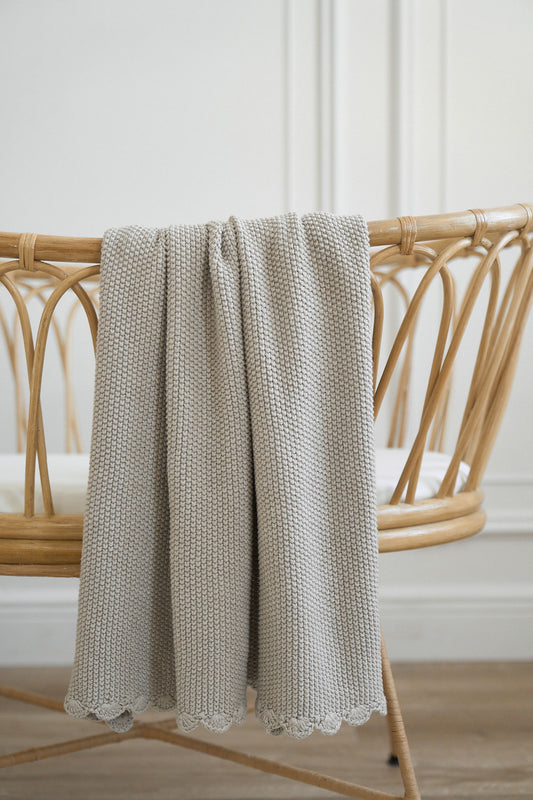 Soft knit mini moss stitch baby blanket in latte color draped over a stylish rattan chair, 100% cotton.
