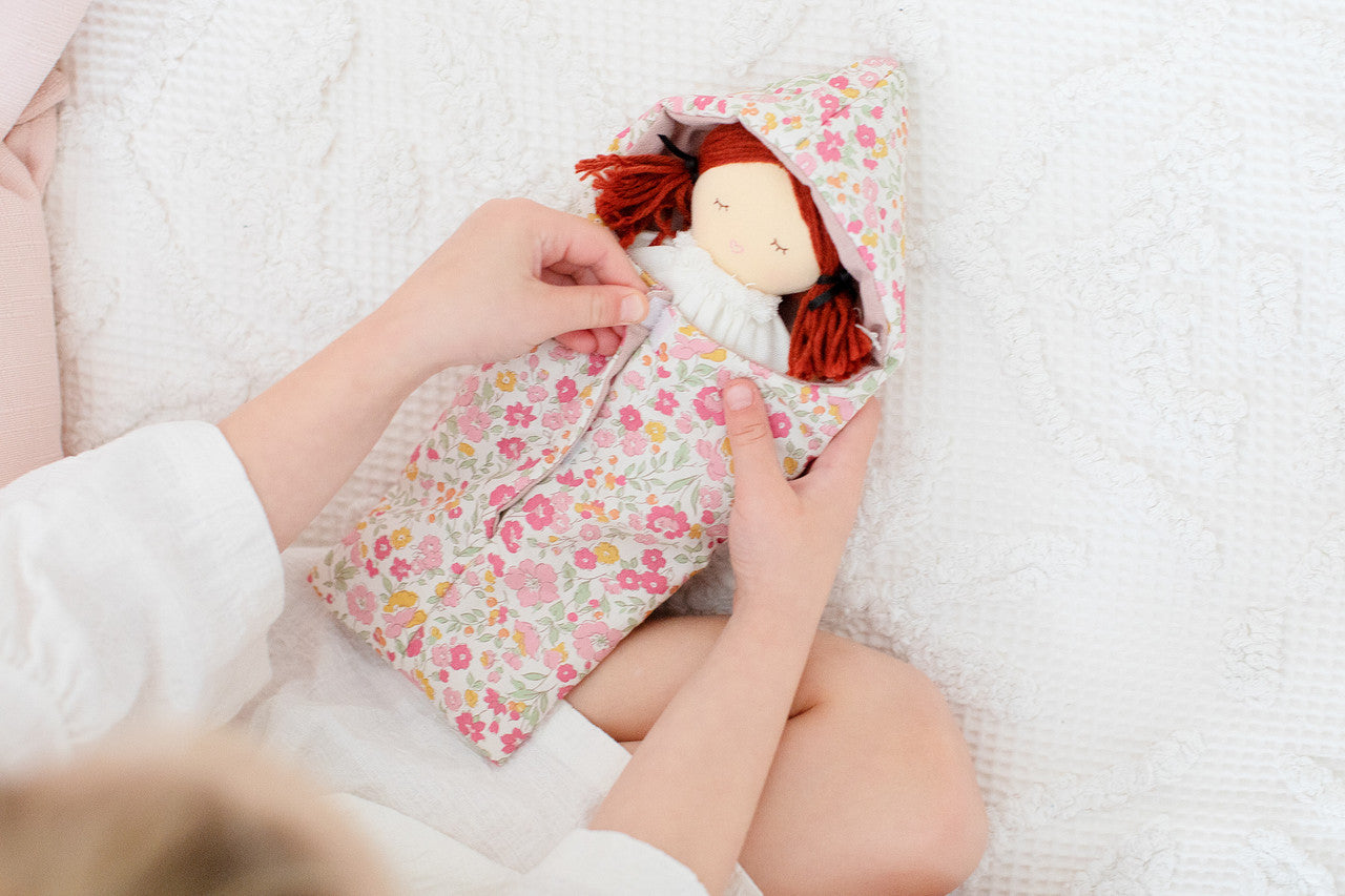A child gently holds the Mini Matilda doll, snuggled in a floral sleeping bag, showcasing its adorable features.