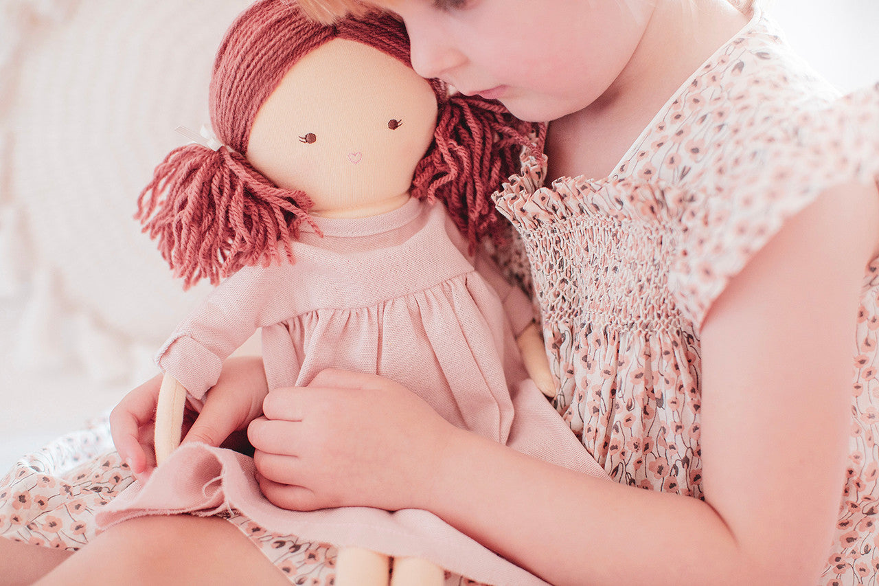 Girl hugging Matilda 45cm doll in pink dress with red yarn hair, perfect for imaginative play and comfort.
