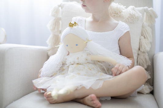 Young girl holding Penelope Princess Doll in gold star tulle dress, sitting on a cozy chair.