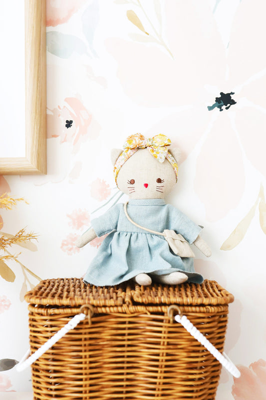 Mini Lilly Kitty doll in grey linen dress and floral headband, placed on a wicker basket against floral wallpaper.