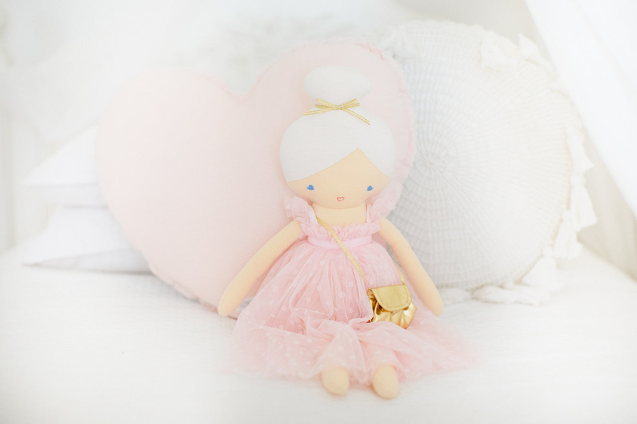 Alimrose Charlotte Doll in pink dress with mini bag, set against a cozy pastel heart pillow.
