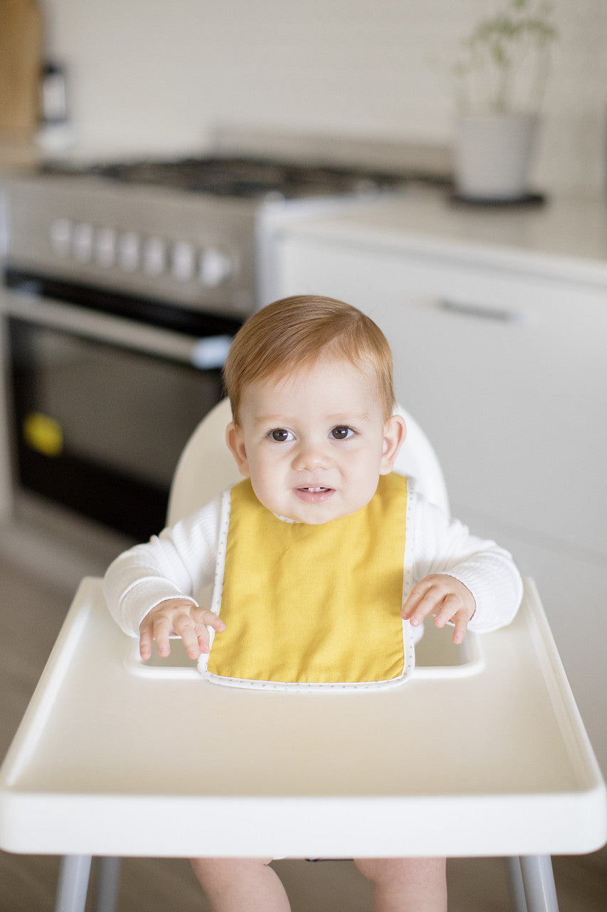 Toddler wearing Bobby Bib Butterscotch Linen, showcasing style and functionality at mealtime in a modern kitchen.