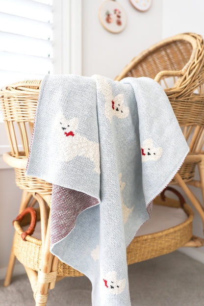 Soft light gray baby blanket with white Westie puppy faces and red bow details draped over wicker chair.