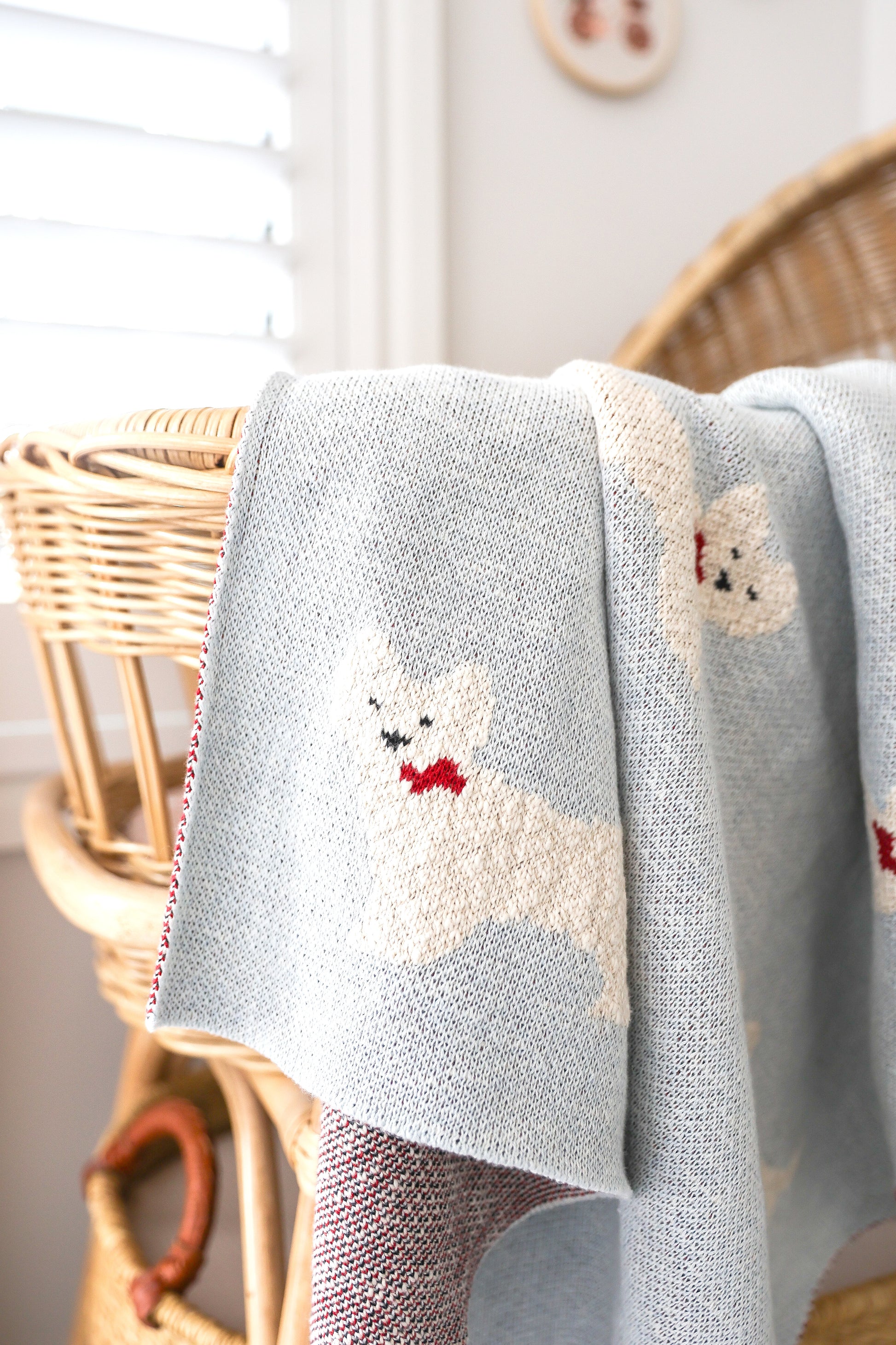 Light blue knitted baby blanket with white Westie puppy and red bow details draped over wicker bassinet.
