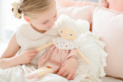 Soft fabric doll with blonde hair, wearing pink floral dress and white lace collar, seated on white cushion.