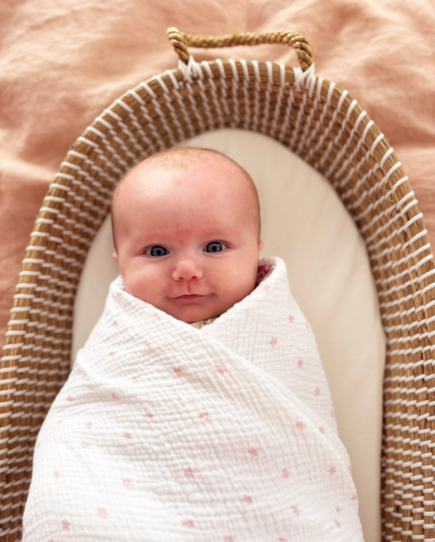 Newborn wrapped in a white muslin swaddle with soft pink star prints in a woven basket cradle.