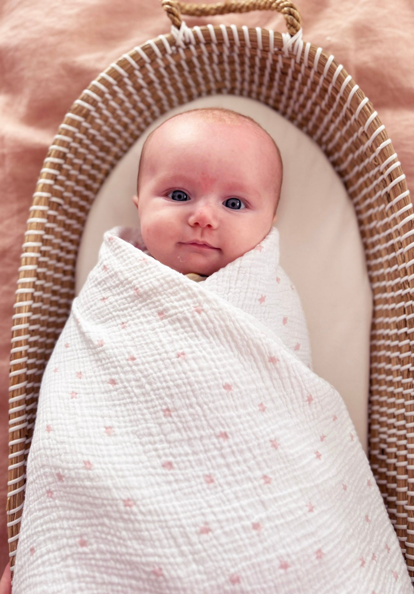 White muslin swaddle with small pink stars wrapped around a baby in a striped cushioned basket.