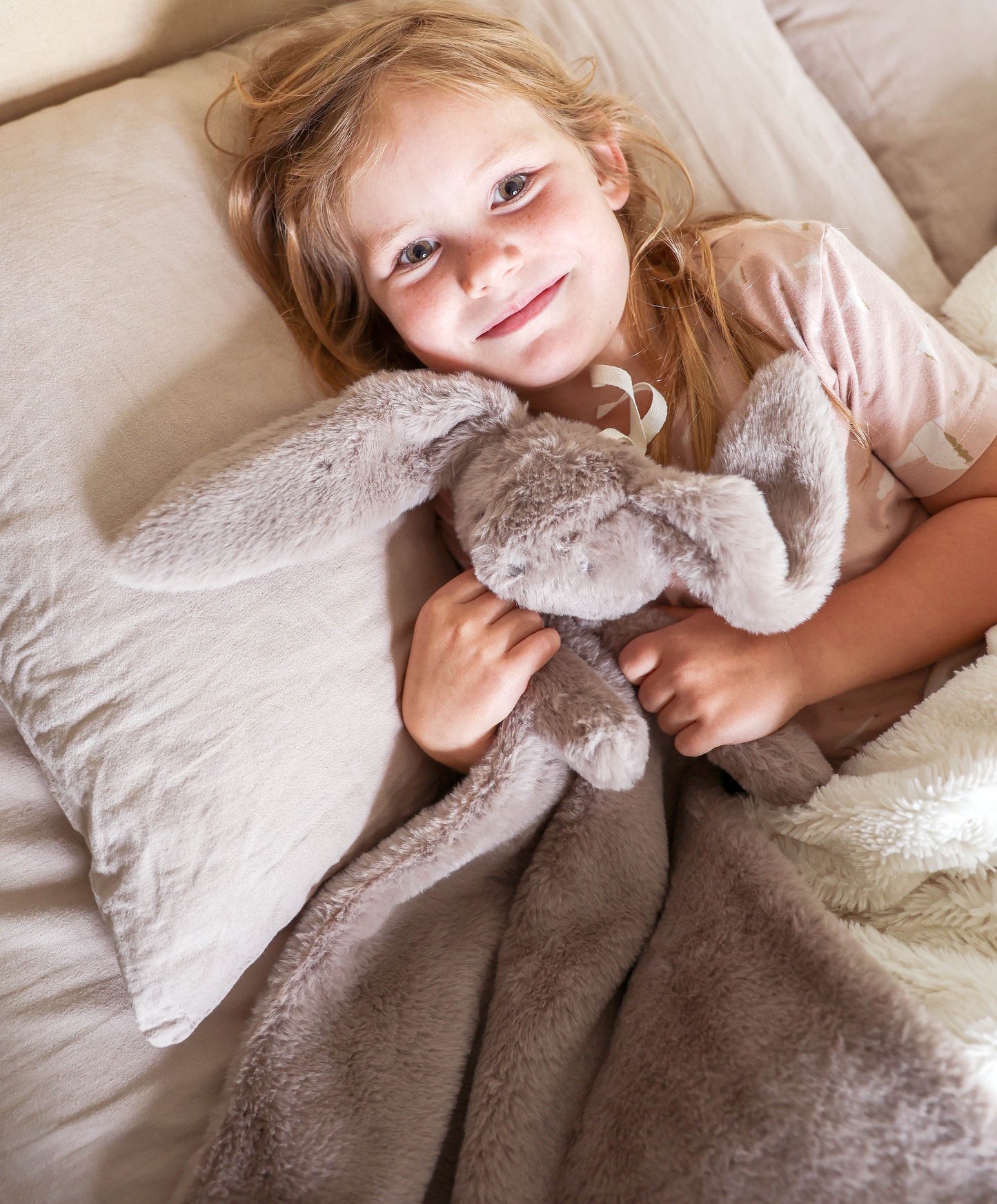 Soft grey plush lovey blanket with floppy-eared bunny attached, held by smiling child in bed.