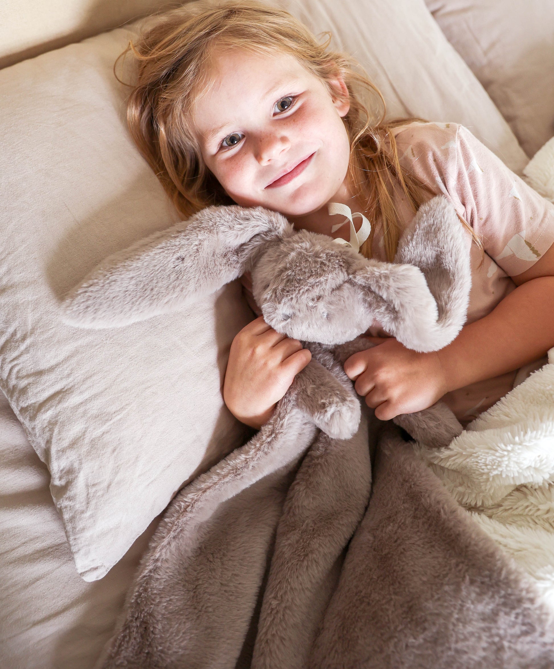 Soft grey plush lovey blanket with floppy-eared bunny attached, held by smiling child in bed.
