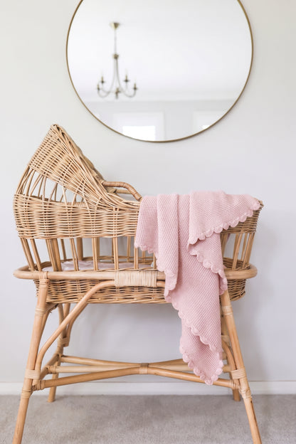 Petal pink knit mini moss stitch baby blanket draped over a natural wicker bassinet with scalloped edges.
