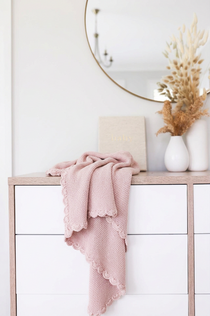 Petal pink knit mini blanket with moss stitch texture and scalloped edges draped over white dresser.