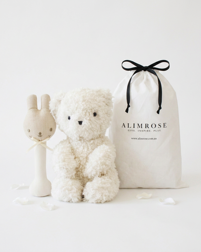 Soft white teddy bear with black eyes and nose, beige bunny rattle with embroidered face, and cream drawstring gift bag with