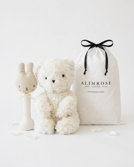 Soft white teddy bear with black eyes and nose, beige bunny rattle with embroidered face, and cream drawstring gift bag with