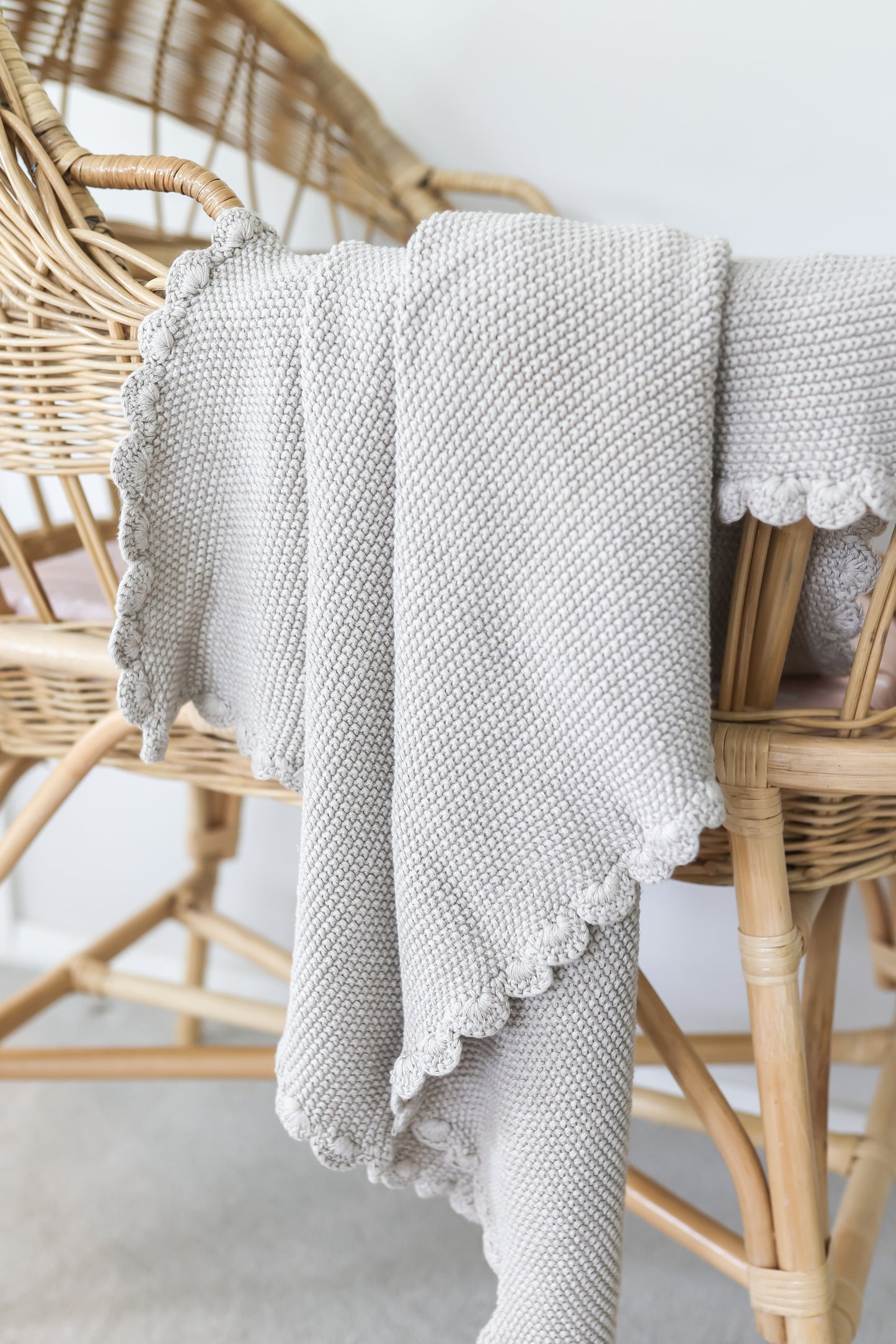 Latte-colored knit baby blanket with moss stitch texture and scalloped edges draped over wicker chair.
