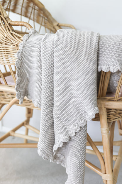 Latte-colored knit baby blanket with moss stitch texture and scalloped edges draped over wicker chair.