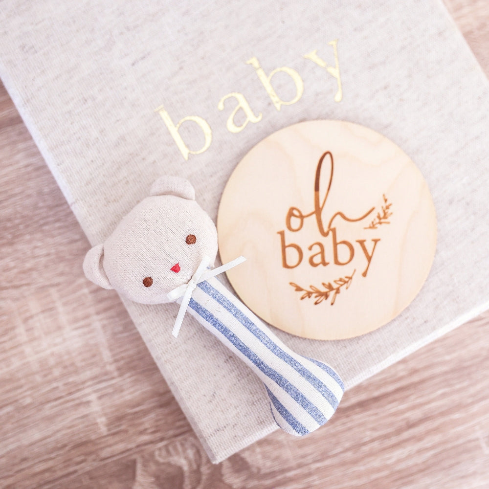 Beige baby blanket with gold "baby" text, striped blue and white plush teddy rattle, wooden disc engraved with "oh baby" and