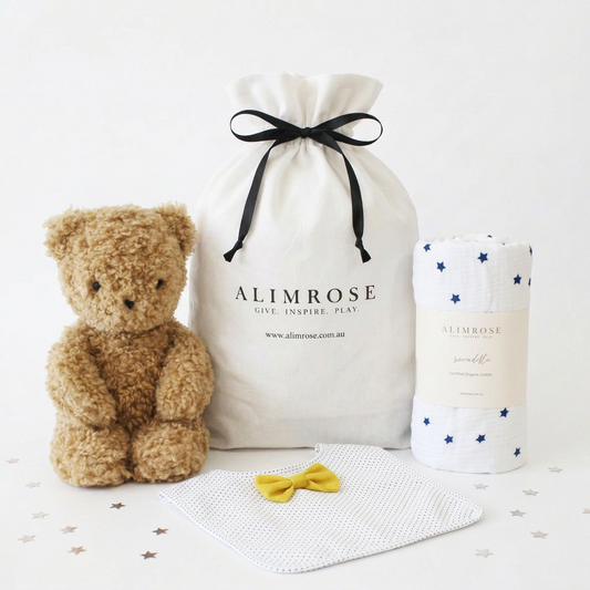 Navy star-patterned baby blanket, soft brown teddy bear, white bandana bib with yellow bow, and cream drawstring gift bag.