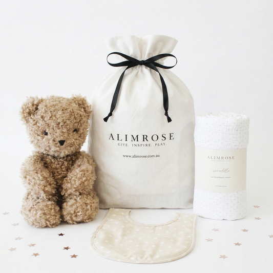 Neutral beige plush teddy bear with a soft drawstring cotton gift bag and white textured baby blanket with subtle pattern.