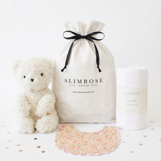 Soft white plush teddy bear, white fabric drawstring gift bag with black ribbon and logo, and rolled white textured baby.