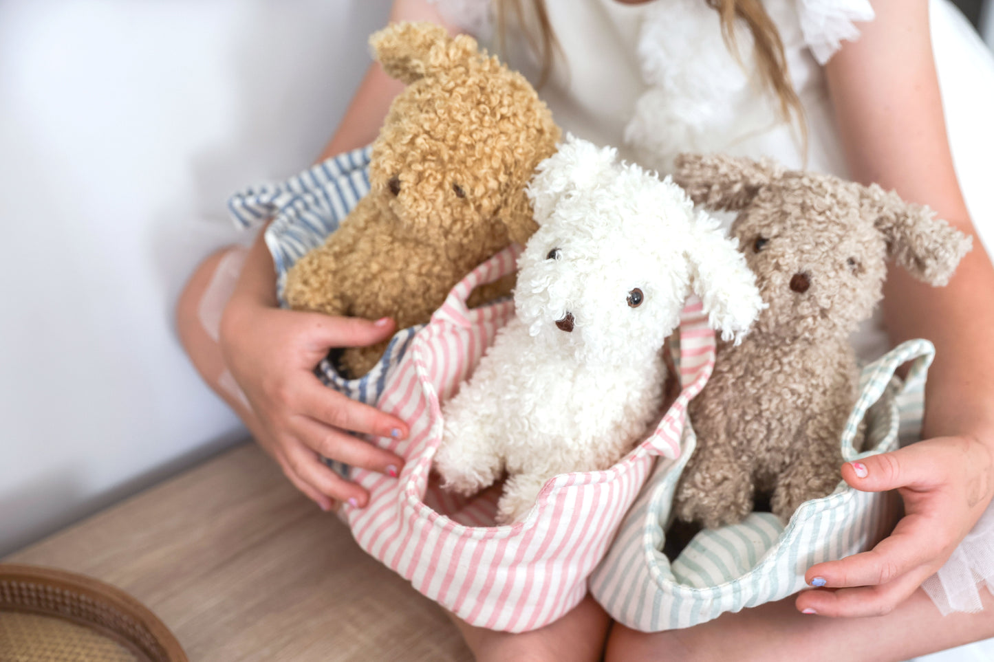 Three plush puppies in light pink and blue striped soft fabric carrier with white trim, held by child with lace sleeve.