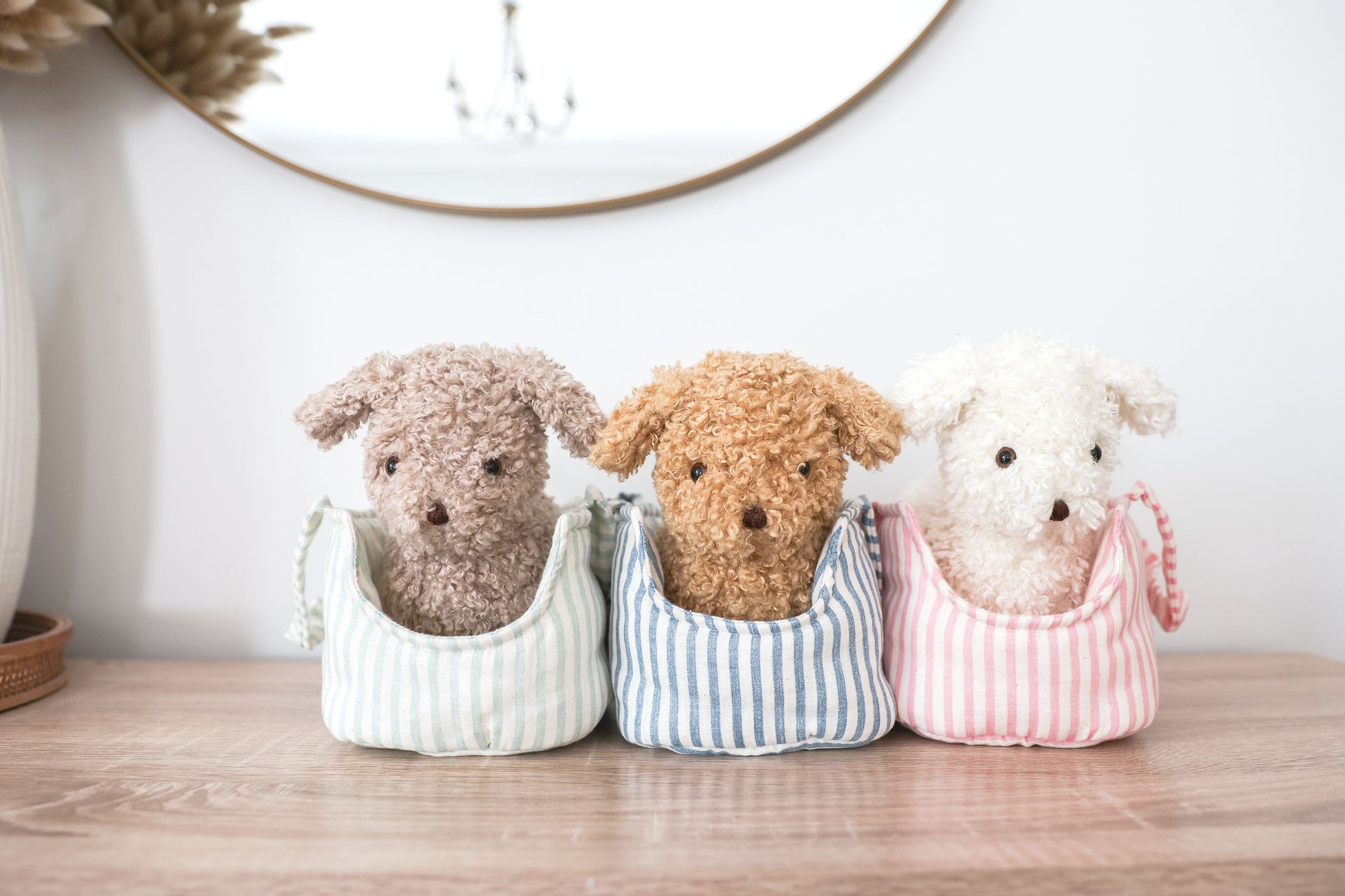 Three plush puppy toys in soft brown, tan, and white, each sitting inside pastel-striped fabric carriers on a wooden surface.