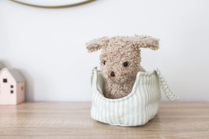 Soft beige plush puppy with floppy ears sitting in light blue and white striped fabric carrier bag on wooden surface.