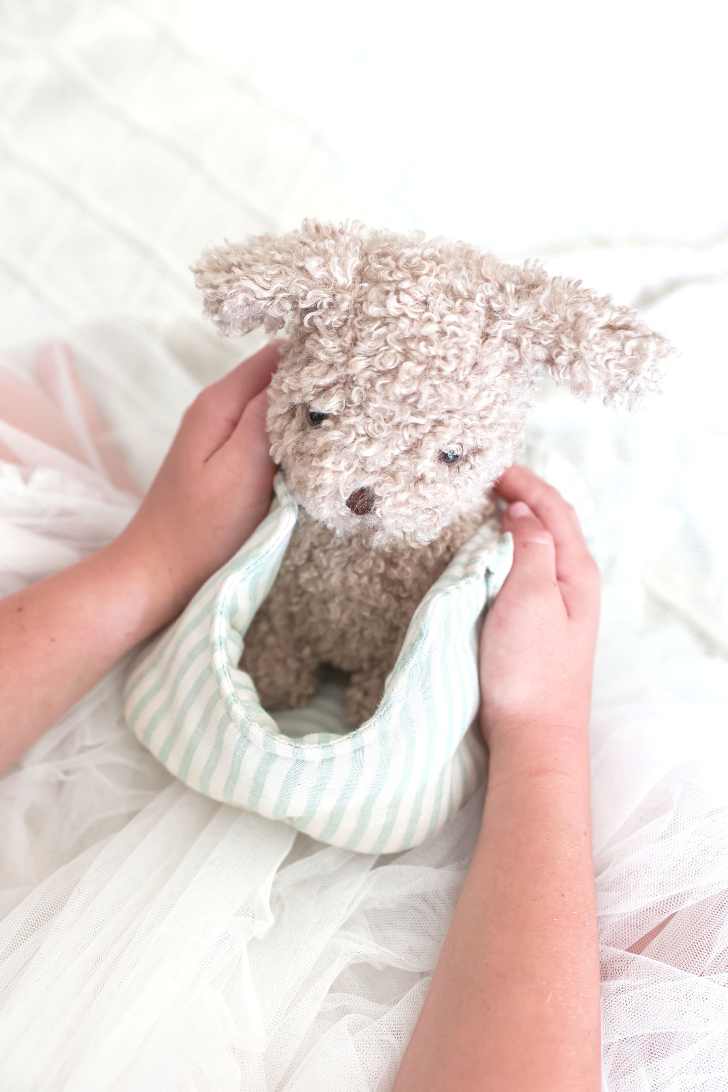 Small beige plush puppy with curly fur nestled inside a soft striped fabric carrier held by two hands.
