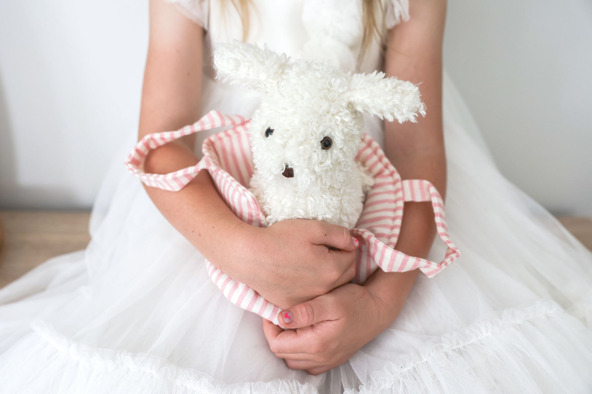 White fluffy plush puppy with black eyes and nose inside a pink and white striped fabric carrier, held by a child in a white
