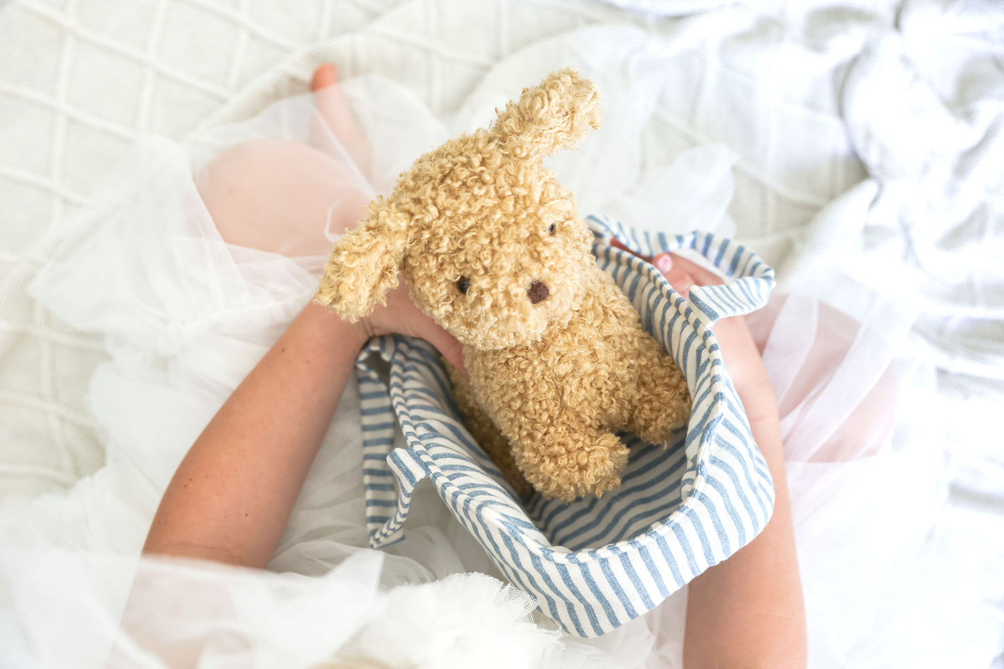 Small curly beige plush puppy named Toffee sitting inside a soft blue and white striped fabric carrier held by child on.