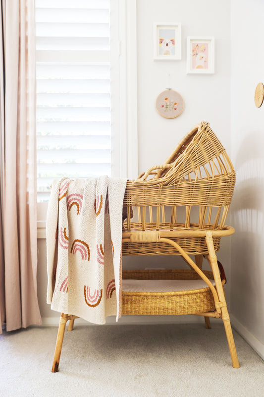 Pink and beige rainbow-patterned baby blanket draped over a wicker bassinet in a nursery.