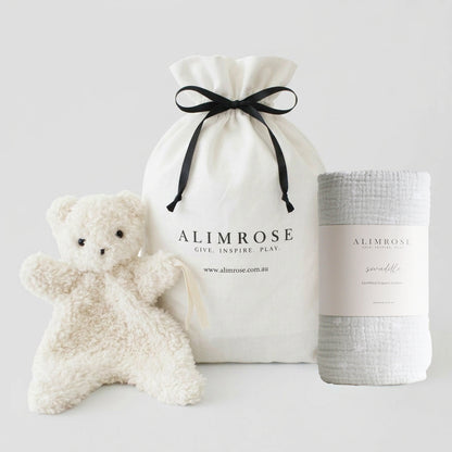 Neutral-colored soft plush teddy bear, white drawstring pouch with black ribbon and logo, and light gray textured blanket.