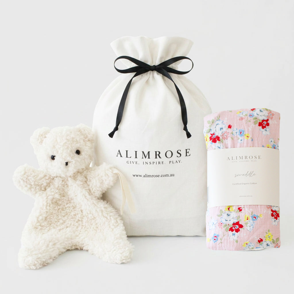 Pink floral print blanket, soft white teddy bear plush, and white drawstring pouch with black ribbon.