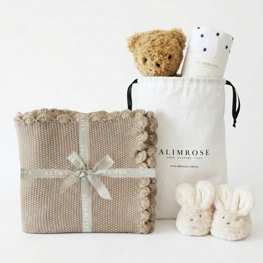 Latte-colored knitted baby blanket with scalloped edges, tied with a cream ribbon, alongside a white branded drawstring bag.