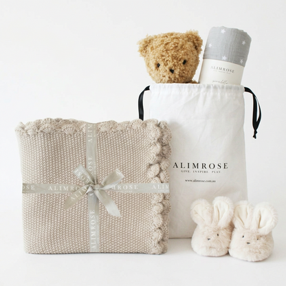 Neutral beige knit baby blanket with scalloped edges, tied with a cream ribbon, next to a white drawstring bag containing a.