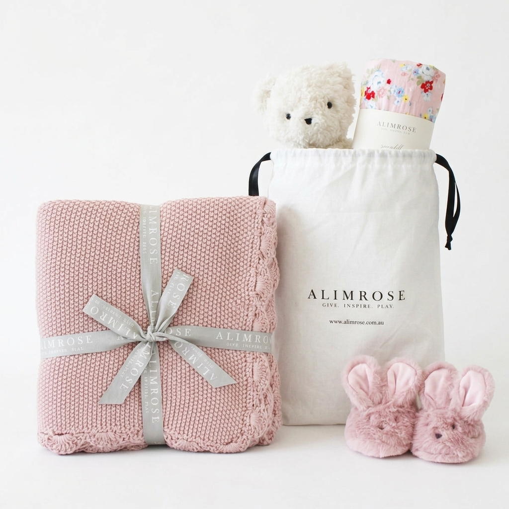 Pink knitted baby blanket tied with silver ribbon, white drawstring bag with plush bear and floral baby wrap, and pink.