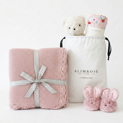 Pink knitted baby blanket tied with silver ribbon, white drawstring bag with plush bear and floral baby wrap, and pink.